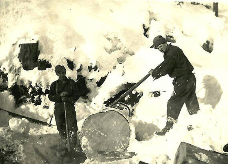 Two CCC men in front of a large pile of logs covered in snow with peavies. Back of the photo reads: 'Using cant hooks to turn the little piece of wood.'