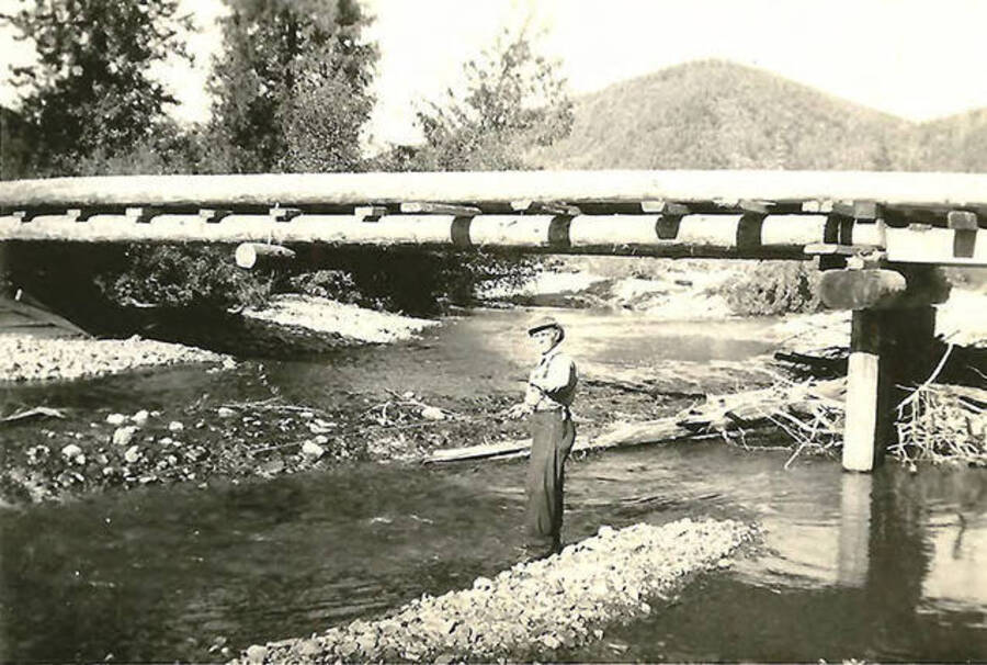 A CCC man stands under the bridge fishing in the creek. Back of the photo reads: 'Another view of bridge over Big Creek. We built them solid.'