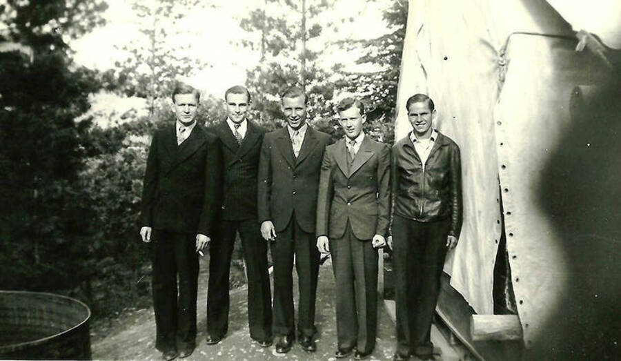 Five CCC men stand in front of a tent barrack at the spike camp in Mullan, Idaho. Back of the photo reads: 'L-R 1st man not ident. 2nd Chick Wethington (Head Cook) 3rd Smokey Gosselt (truck driver) 4th man not ident. 5th Ed Sash (from near Steubenville)'.
