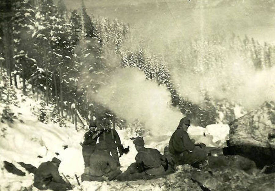 Six CCC men sitting in the snow in the middle of the woods around a camp fire. Snowy woods can be seen through the smoke. Back of the photo reads: 'Snow bunnies at work? Probably lunch time. We carried 2 sandwiches and on apple in a cloth 5 lb sugar sack tied to our belt.'