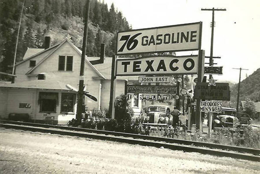 A view of railroad tracks going past a 76 Gasoline station as the top sign advertises. Other signs read: 'Texaco' 'John East' 'Insulated' 'Star Brano Shoes' 'Theodore's Men's Wear'. Back of the photo reads: 'Train tracks thru Burke, Idaho - Mining town near Mullan. They go right down the main street.'