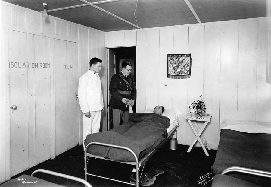 Two men visit a sick man in the Isolation Room at CCC Camp SCS-1 near Moscow, Idaho. The signs on the wall read: 'Isolation Room' and 'Men'. The small tapestry on the wall reads 'mother'.