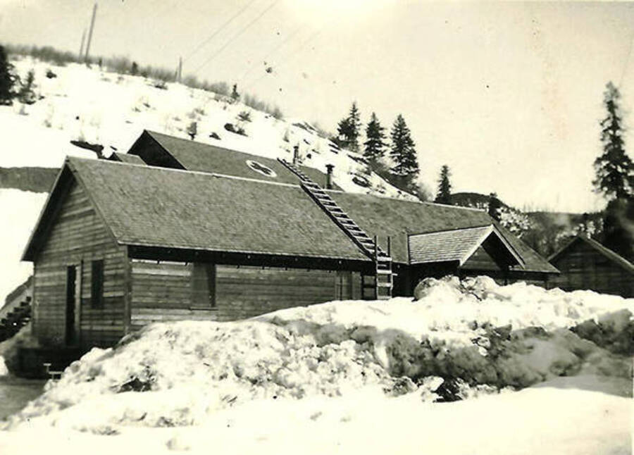 A view of the Infirmary at CCC Camp Big Creek #2, F-132. There is a pile of snow in front of the building and a hill rising in the background. The sign of the red cross is on the roof. Back of the photo reads: 'winter of 1939. We had plenty of snow to play in.'