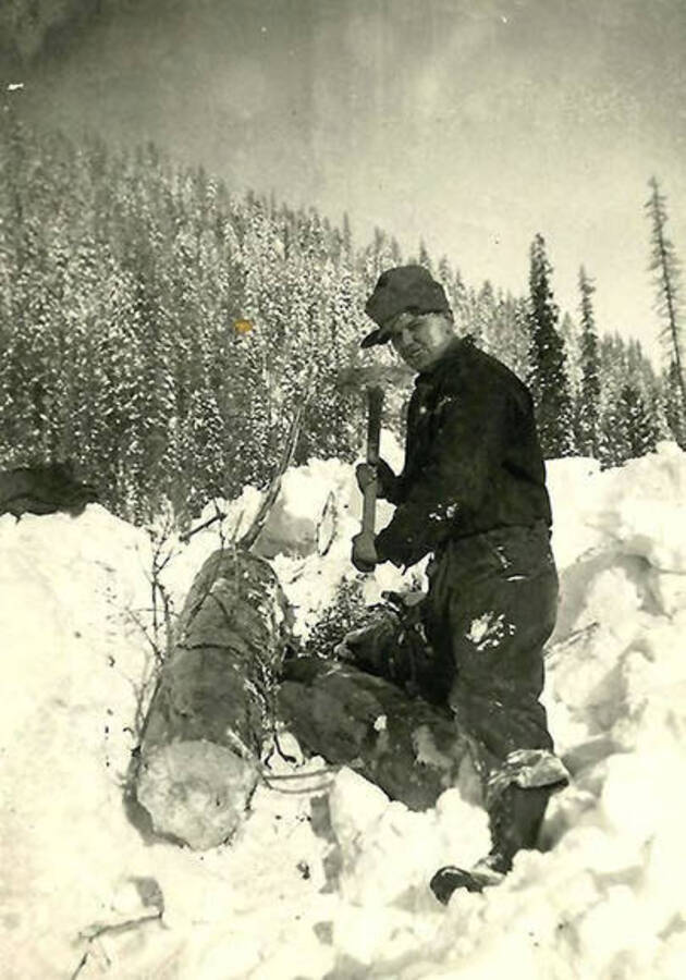 A CCC man sits in the snow holding an ax. He is sitting next to a log outside of CCC Camp Big Creek #2, F-132. A forested hill rises in the background. Back of the photo reads: 'Getting out the logs we used for cribbing up the edge of the roads we built. All dirt in those days.'