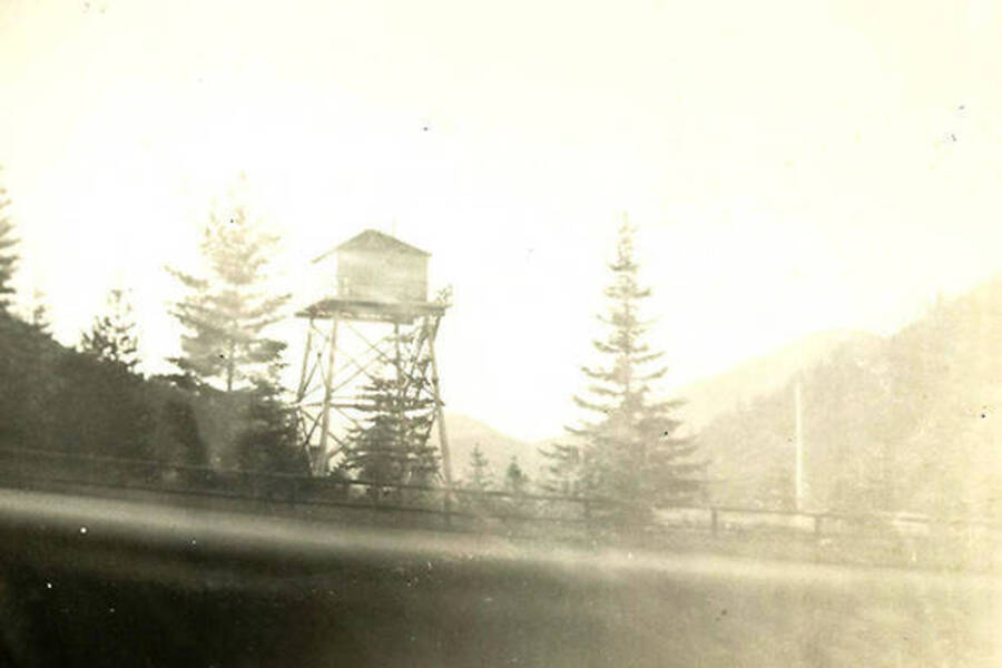 A lookout tower can be seen among the trees. A fence lines a ridge below the tower. Back of photo reads: 'Fire tower near Mullan Pass'.