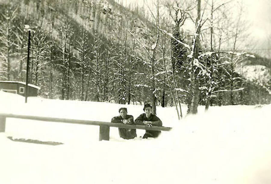 Two CCC men lean against a fence that disappears underneath a pile of snow at CCC Camp Big Creek CCC Camp #2, F-132. Behind them is a snow-covered field, forest and hill. Back of the photo reads: 'near Camp Big Creek, (1939 winter).'