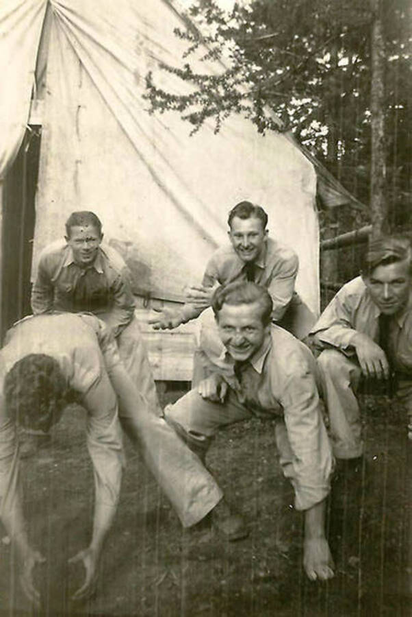 Five CCC men pose Like linemen and the quarterback in front of a tent barrack at the Mullan, Idaho spike camp. Back of the photo reads: 'Our team, we had fun'.