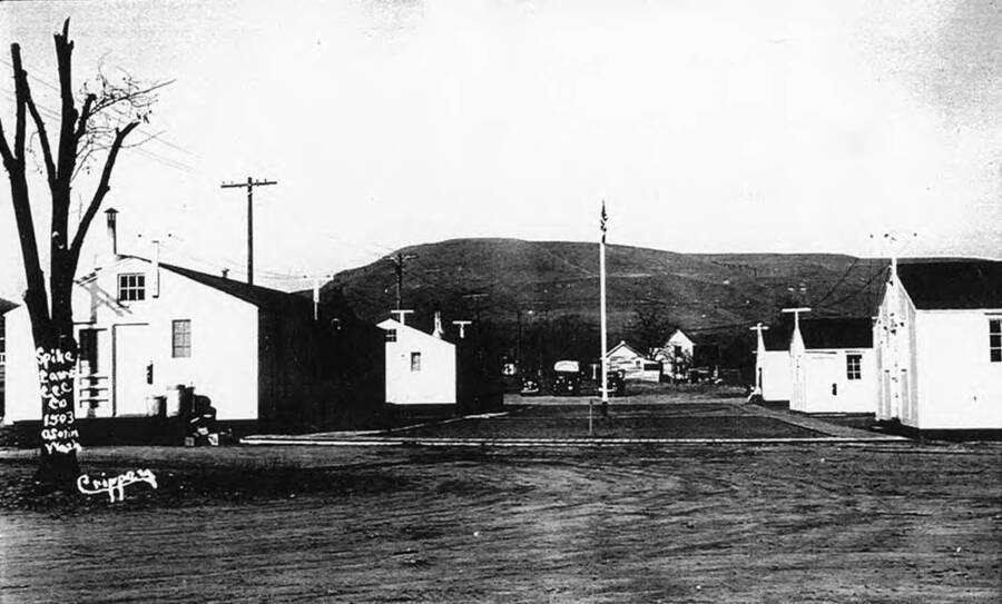 CCC Spike Camp in Asotin, Washington. There is a flagpole in the middle of a courtyard that is surrounded by five white buildings. A dead tree sits in the foreground of the photo and a few trucks and buildings can be seen in the background. Writing on the photo reads: 'Spike Camp CCC Co 1503 Asotin, Wash. Crippen'