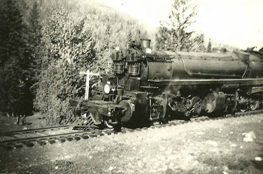 A train engine sits on the railroad tracks in the woods. Back of the photo reads: 'Close up of engine on Northern Pacific line near Mullan. Note size of engine for hauling freight thru mountains.'
