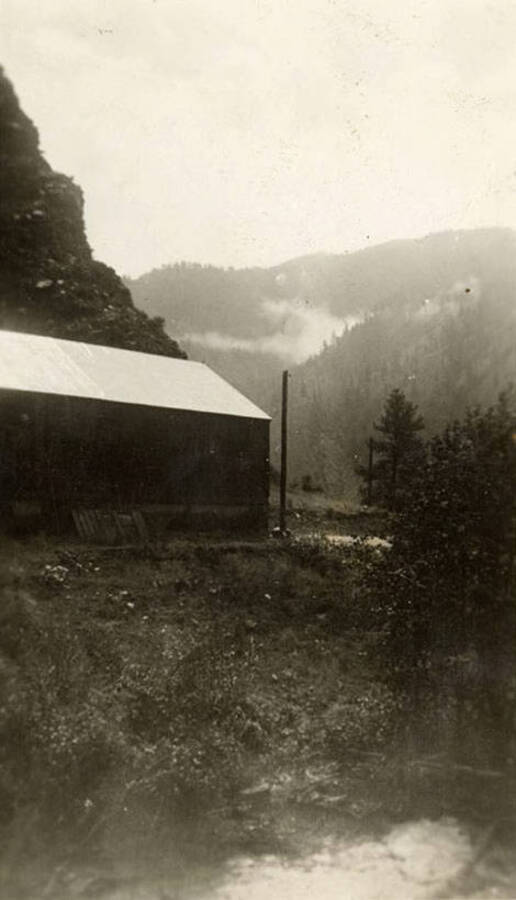 A CCC building sits just above the Salmon River, with hills rising steeply in the background. Back of the photo reads: 'Camp Cove Creek F-401 1937/38 Salmon River right below our Co. 2763 Admin Office my bedroom was right at the left side of this picture.'