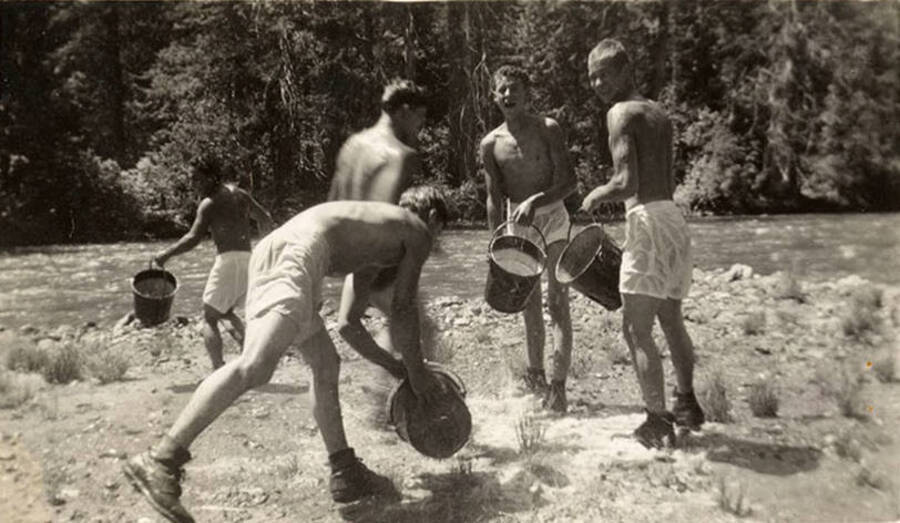 Five CCC men in white swim tunks and boots holding buckets full of water next to a river. Back of the photo reads: 'Water fight - main camp August 1938'