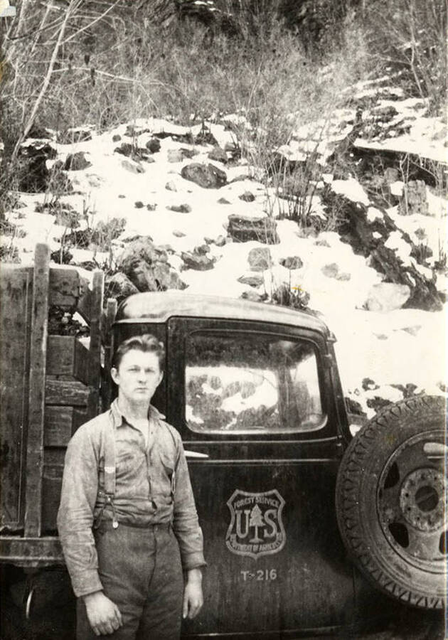 A CCC man stands in front of a Forest Service Truck, which is parked below a snow-covered slope. The seal on the truck reads: 'US Forest Service Department of Agriculture' and the label below the seal is: 'T-216' Back of the photo reads: '1938-1939- Aver, Idaho CCC - Herb with his blond wavy hair - hauling dynamite for the Fishhook Creek Tunnel Company 1239, F-187 Avery, Idaho. A company also along the St. Joe River in the Bitter Root Mts.'