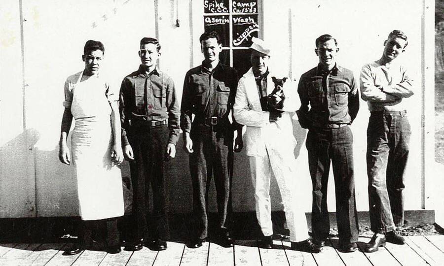 Six uniformed CCC men stand outside on a porch in front of a building. One of the men wears a cooks apron, another a white suit and hat. The man with the hat is holding a puppy. The writing on the photo reads: 'Spike Camp CCC Co. 1563 Asotin, Wash. Crippen.'
