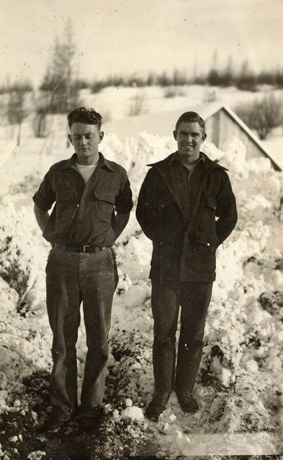 Two uniformed CCC men standing in front of a large pile of snow, obscuring a snow-covered building and field in the background.
