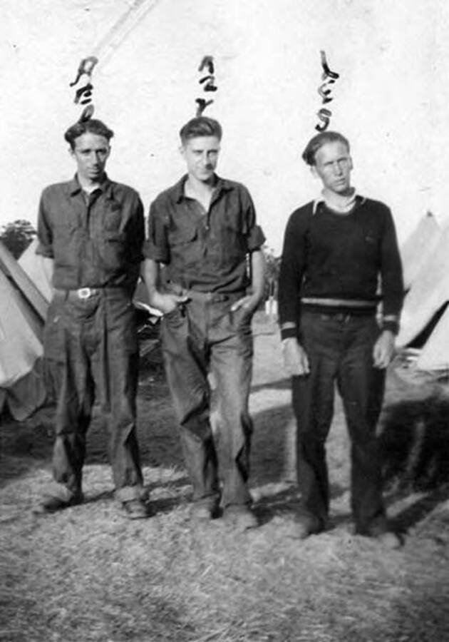 Three CCC men standing in a CCC tent camp. Above each of their heads their names have been written. They read left to right: 'Red, Azy, Les' and below the picture reads: 'Part of gang'.