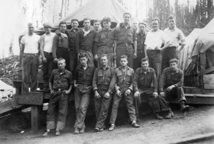 Group photo in front of a tent at the CCC camp. Most men are in uniform but two enrollees on each end are wearing white t-shirts. Writing over the photo reads: 'Company 1290 CCC Idaho 1933'.