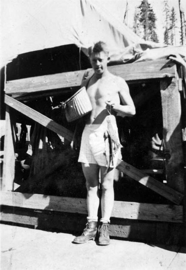 A CCC boy stands in his shorts carring a basket and a string of fish. He is standing in front of a wooden framed structure that is covered by canvas.
