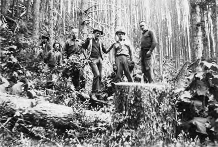 Several CCC Foremen standing behind a downed tree in the forest. Writing on the photo reads: 'Our Foremen'. Writing below the photo reads: Sharker Schk, Red-Schen, All Gornsey Idaho Micky-Troy'.