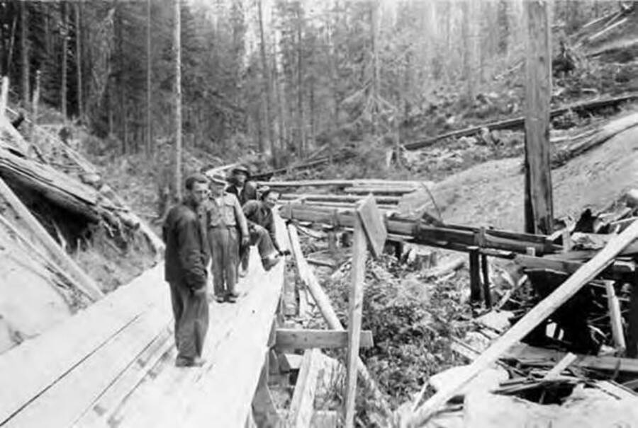 Several CCC men standing in a flume  with other flumes nearby either under construction, or deteriorating. Writing next to the photo reads: 'Gang leaving for work'.