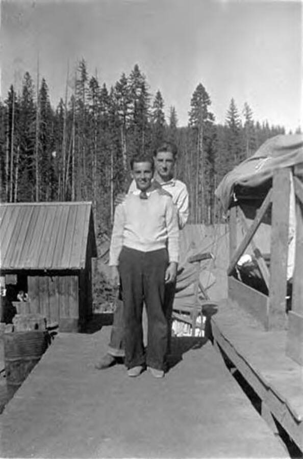 Two CCC men posing on a wooden structure with a building and forest in the background. Writing next to the photo reads: 'Bill - Searight = 020 Ray Saladin'.