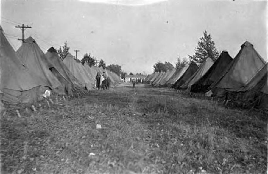 Several men walk through a row of tents in a CCC camp. Writing next to the photo reads: 'Co. ST. Camp Dix 18th Discharge'.