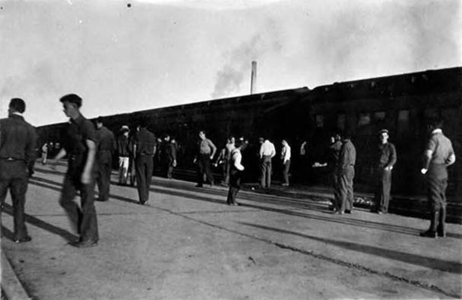 Several CCC men waiting around on a train platform and exiting the train. Writing next to the photo reads: 'Train from the West'.