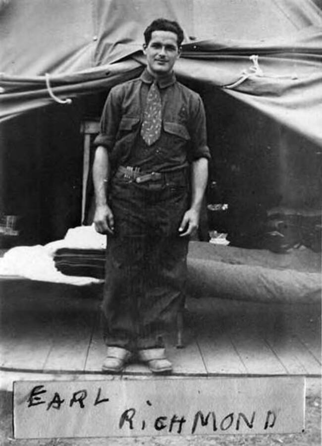 A CCC man stands in a tent in a CCC Camp. There is a cot in the background.Writing on the photo reads: 'Earl Richmond'.