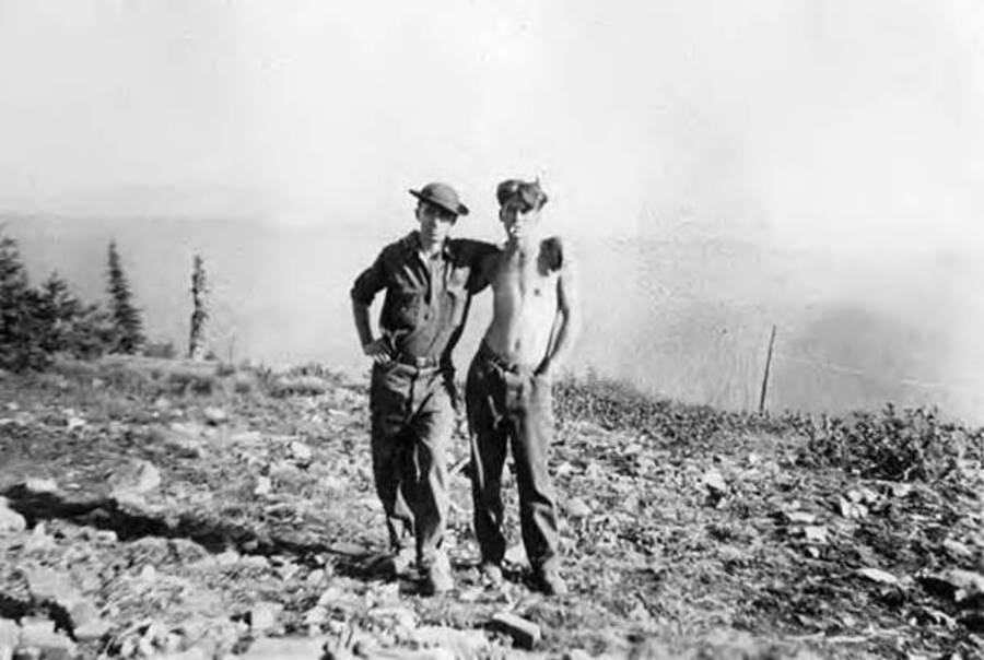 Two CCC men standing and posing for a photo in front of a faint landscape. The man on the right  is posing shirtless with a cigarette in his mouth. Writing below the photo reads: 'Redmond Bohanan'.