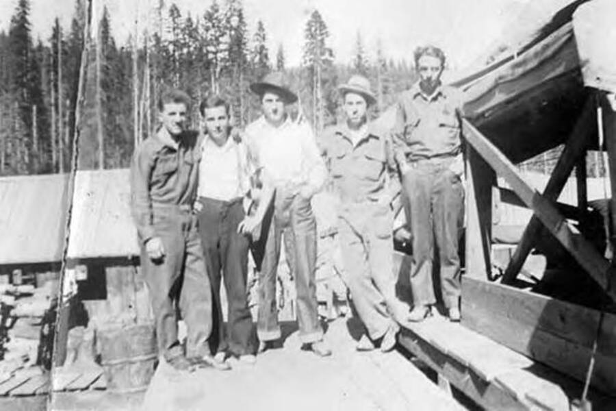 Five CCC men posing for a photo on a wooden structure with a building and forest in the background. Writing under the photo reads: 'Joan - sch. S. Right - Ogo Rhy - Whall J Frisslet & Sulz Red Watll'.