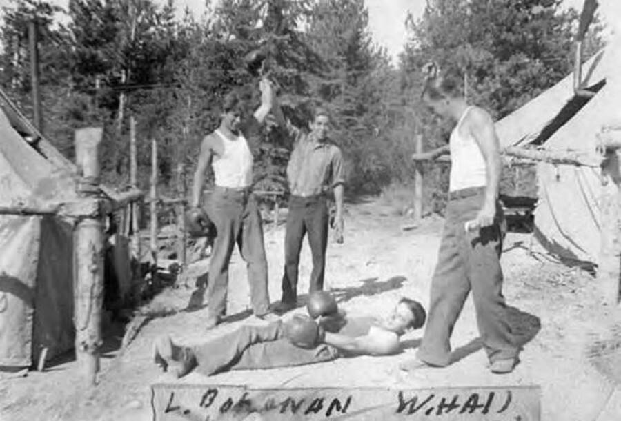 Four CCC men posing for a photo in a CCC camp. Two of the men have boxing gloves on, one is on the ground, while the other has his fist raised in the air. Writing under the photo reads: 'L. Bohanan W.Hall C. Lesett W.Hall P. Gordon W.Hall C. Roberts Granum'.