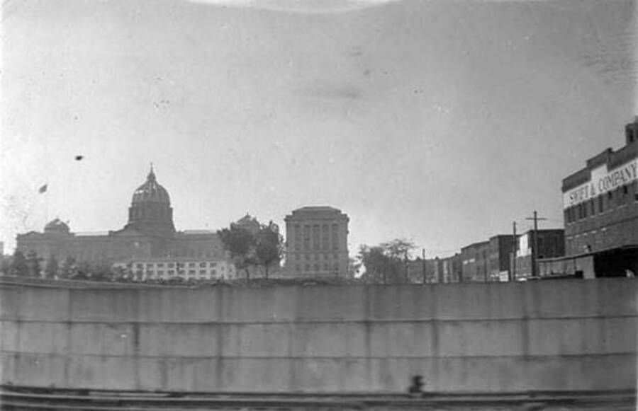 Photo taken from the train of Harrisburg. Above a short concrete wall are several buildings. One advertizes 'Swift & Company'. Writing under the photo reads: 'Harrisburg, Penn'.