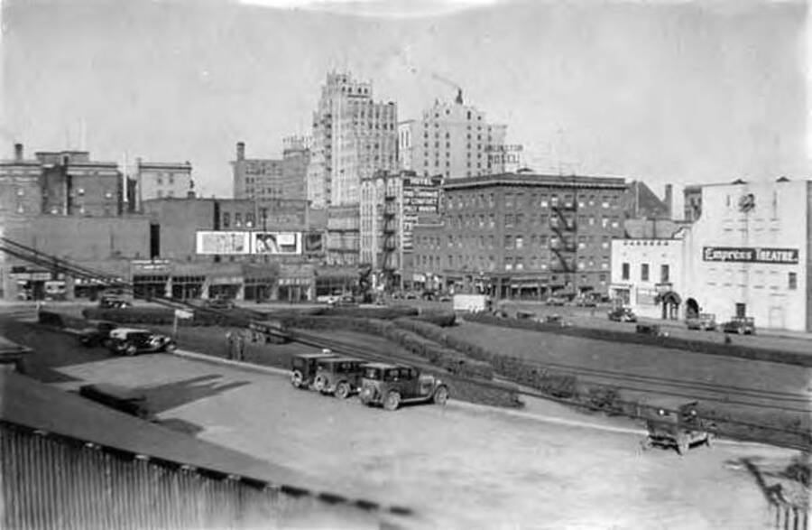 Several industrial buildings can be seen from the railroad station in Spokane, WA. There is also a theatre 'Empress Theatre' as well as a parking lot and some landscaping in the foreground. Writing under the photo reads: '[Spokan]e, [Wa]shington'.