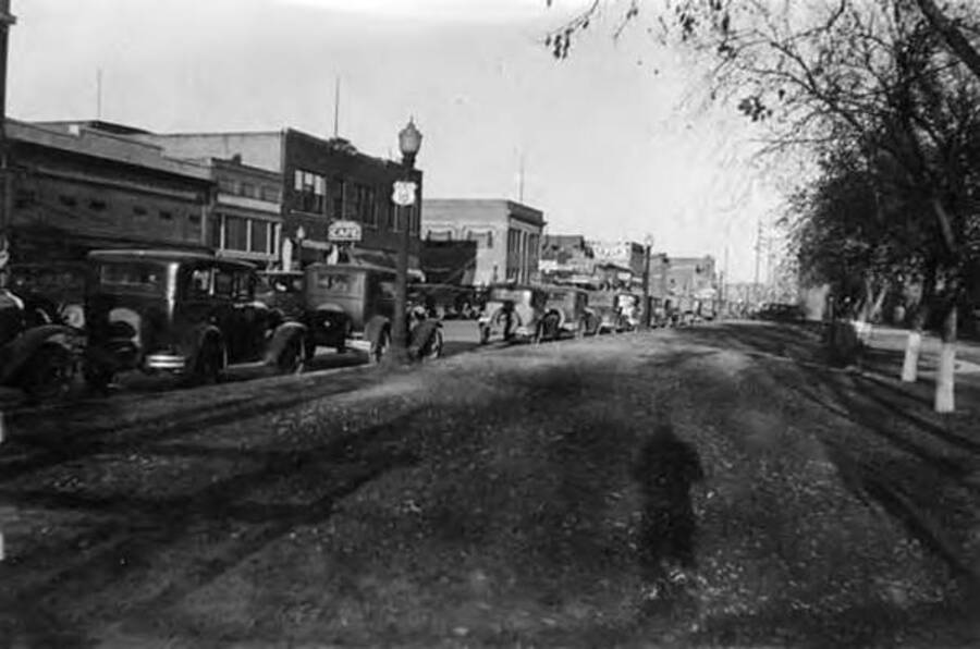 A street lined with trucks in the morning light. The photographer's shadow can be seen in the foreground. Two signs read: 'Cafe' and 'U.S. 10' Writing next to the photo reads: 'Sidney Nebraska'