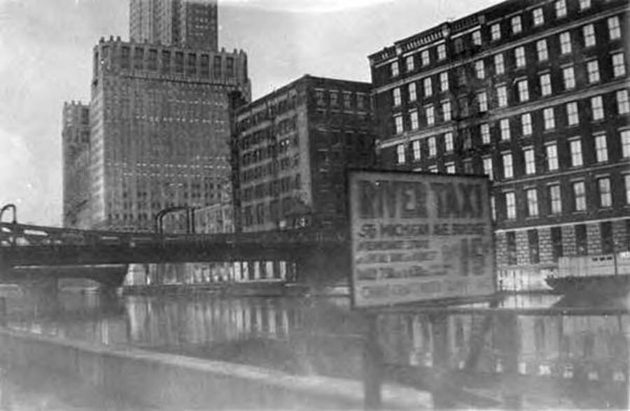 Chicago, IL seen from the train, with a river, bridge, and sign in the foreground, and buildings in the background. Writing on the sign says: 'River Taxi 56 Michigan Ave. Bridge, 15'.
