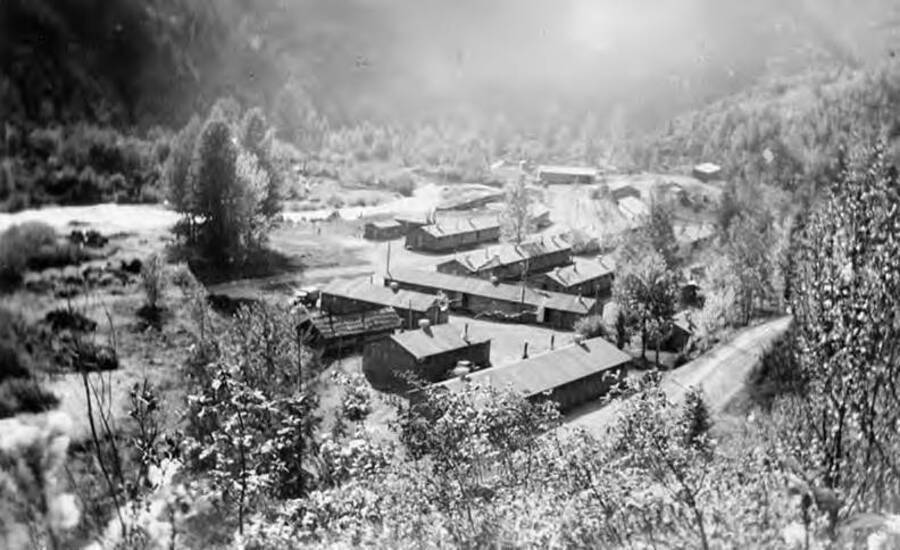 A photo taken from the hill above a CCC camp. Several buildings lay at the base of a valley. A road and creek can be seen meandering through the valley floor. Trees surround the camp.