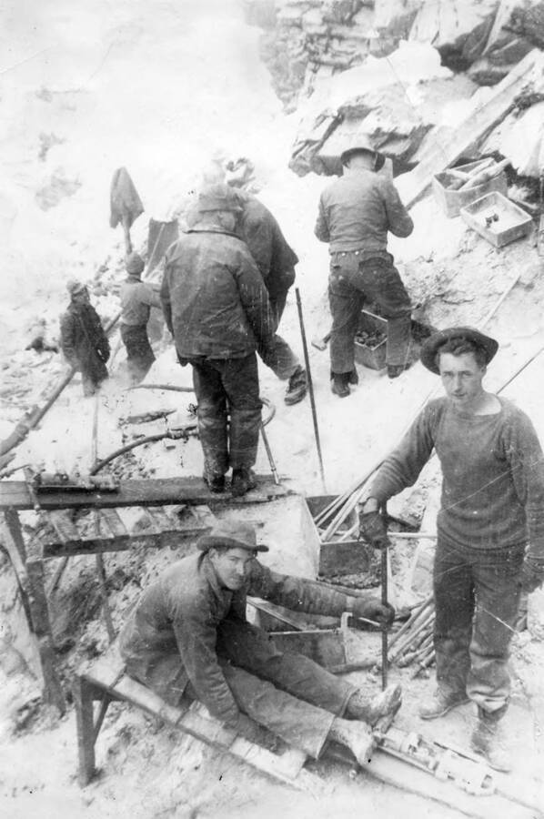 Several CCC men at work in a rocky environment. The photo is taken from above and two of the men working are posing for the camera. Tools and various supplies are strewn about the worksite, some of them in boxes.