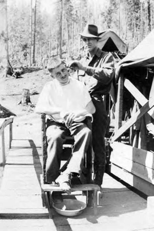 Two men in a CCC camp. One is sitting in a chair, wearing a small smock, and the other man is standing next to him holding a razor or a pair of siccors to the first man's head. Writing below the photo reads: 'Red DeLarens O'Russel Vitalo Bear Our Company Barber'.