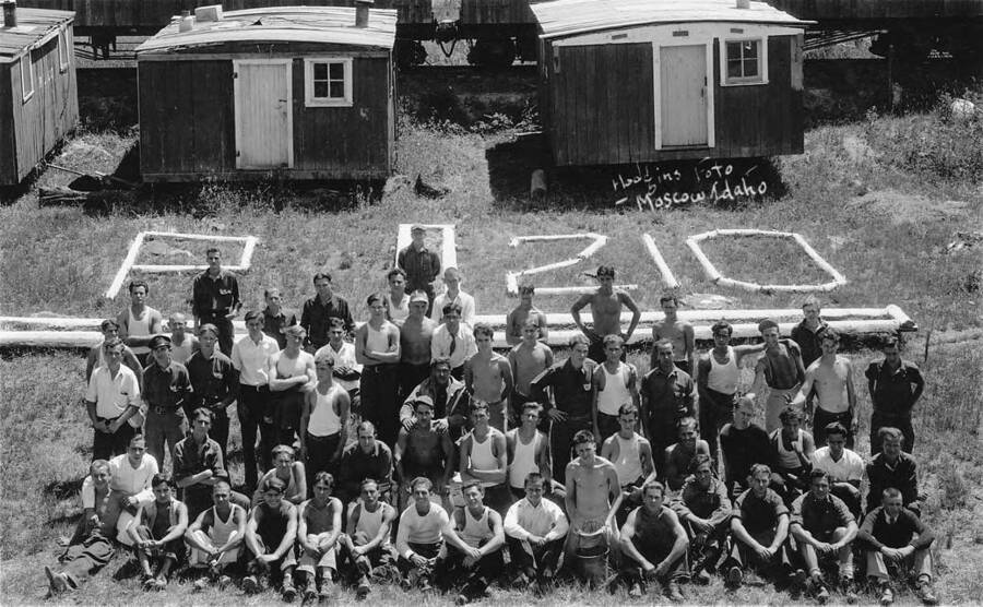 Group portrait of company 1279 at camp P-210 near Bovill, Idaho. Logs laid out read: P-210. Writing on photo reads 'Hodgins Foto - Moscow Idaho'.