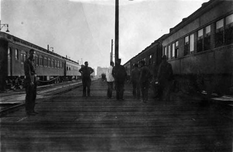 A view of the train platforms at the Elgin, IL train station. Several CCC men stand on the platform and two trains can be seen on either side.