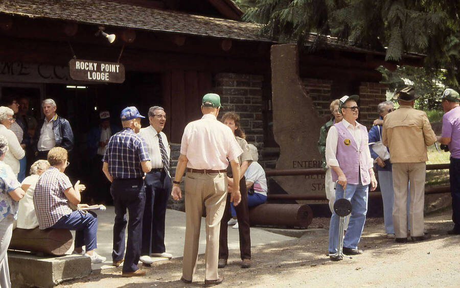 Several elderly men and women outside of the Rocky Point Lodge and the entrance sign which reads: 'Enterin[g] Heyb[urn] Sta[te] [Park]'. One of the women in the center of the photo is wearing a Forest Ranger uniform. The sign behind the lodge sign presumably reads: '[Welco]me C.C.[C.]'.