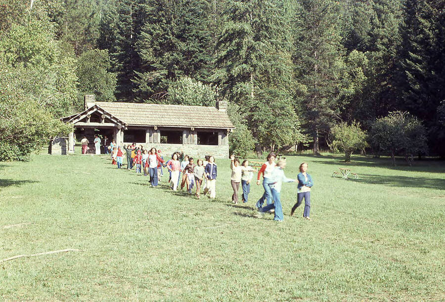 A line of children can be seen moving along the grass away from the Chatcolet Shelter at Heyburn State Park. Several picnic benches are scattered across the lawn behind them.