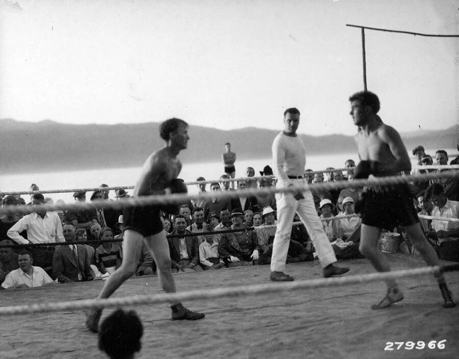 Two boxers circle each other in a boxing ring observed by a referee and an audience. Back of photo reads: 'ECW Historical Record 279966 Boxing wound up the day's sports at the CCC carnival at McCall, Idaho. Three camps competing. Near Idaho National Forest. Taken by K.D. Swan.'