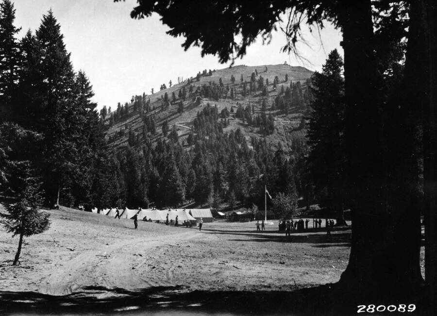 CCC men standing in a field (they may or may not be playing baseball) in front of their CCC Camp and a flag pole, which is in front of a large wooded hill.