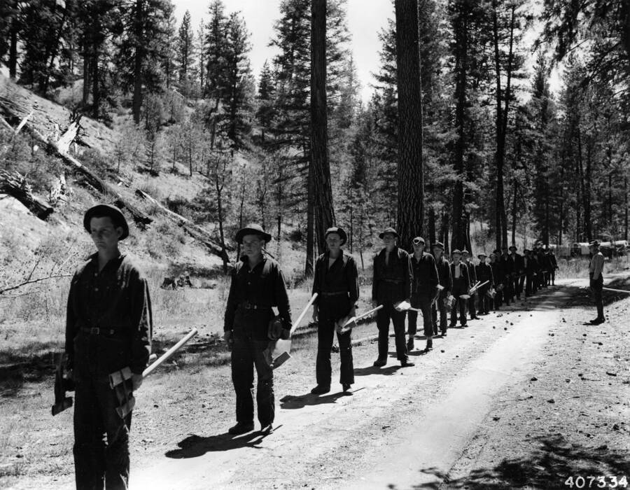 A CCC work crew stands in a line on a dirt road in the wood. Each enrollee is holding an axe with a covered edge. An officer stands at attention on the other side of the road. In the distant background is a parked row of covered trucks.