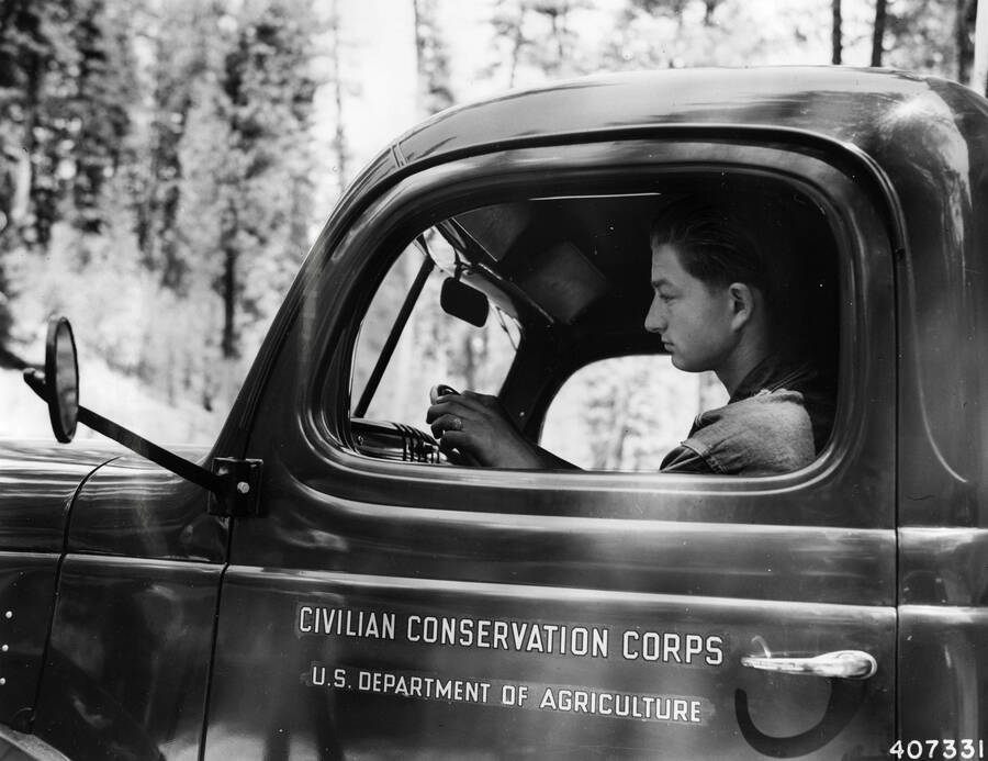 A CCC man sits at the wheel of a truck. On the driver's side door there is lettering that reads: 'Civilian Conservation Corps U.S. Department of Agriculture'.