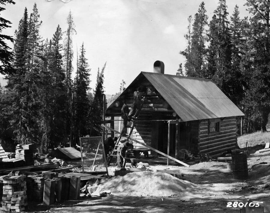 Several men working on a cabin, a forest sourrounds them and a multitude of construction supplies sits in front of the cabin.