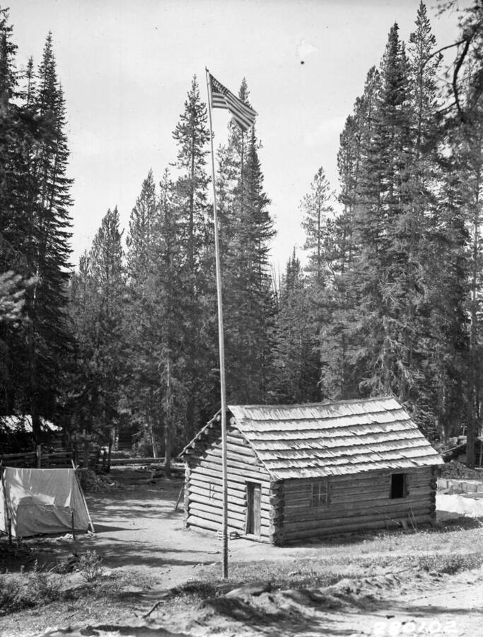 Photo of a flagpole in front of a cabin in the woods. A small tent sits to the side of the cabin.
