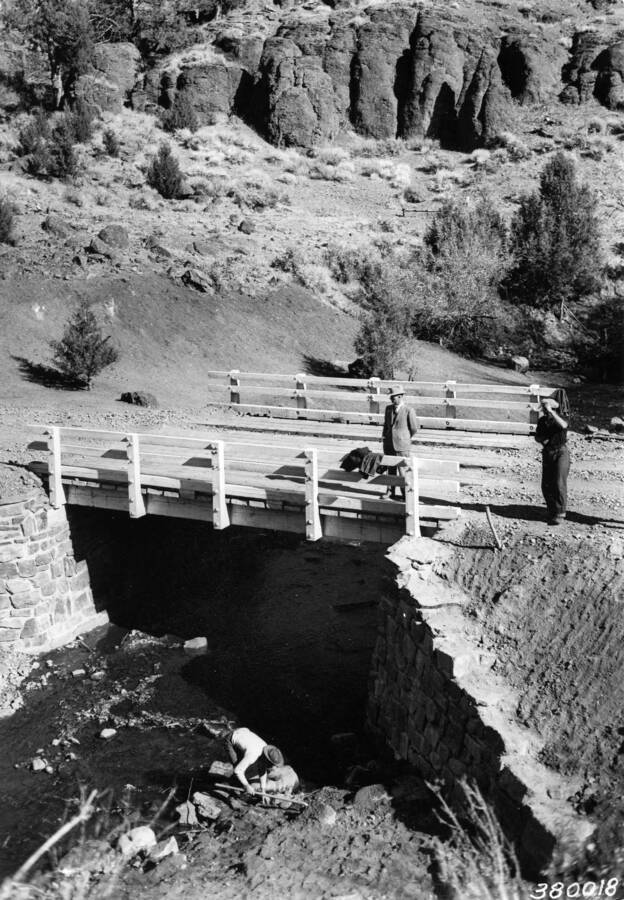 Two men standing above a bridge and one man below working on the bank of a creek. The bridge may have been constructed by the CCC.