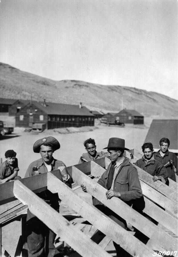A CCC work crew constructing a building a a CCC Camp. Barrack buildings and hills appear in the background.