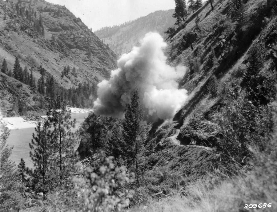 An explosion in the Salmon River Canyon on the Salmon River road project. Description reads: 'Shooting rock points on Salmon River road above French Creek. A CCC job. Forest: Nez Perce, State: Idaho, Date: 7/1935, Author: K.D. Swan'.